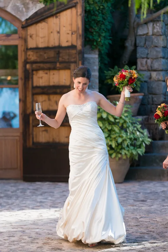 Smiling bride in a strapless white wedding gown holding a bouquet and a champagne glass outdoors.