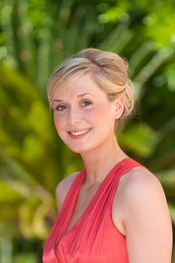 Smiling woman with blonde hair styled in an updo, wearing a sleeveless coral dress, standing outdoors with blurred green foliage background.
