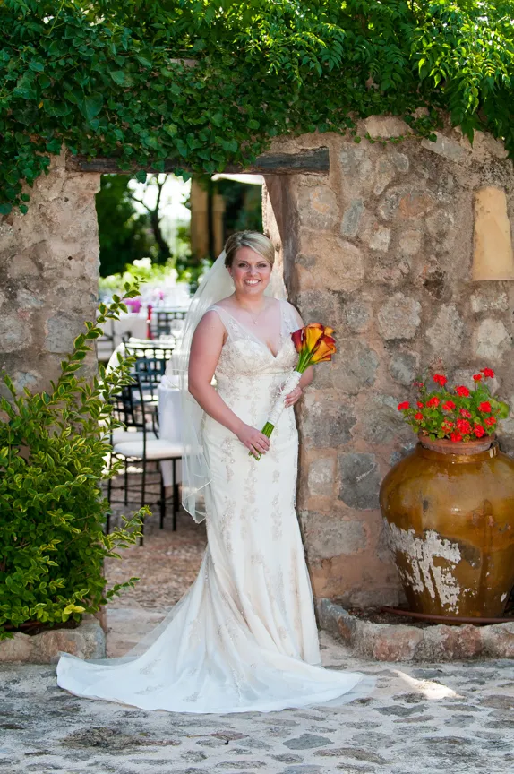 Bride in a white lace wedding dress holding orange calla lilies, standing in a rustic stone archway with greenery and a large pot of red flowers.