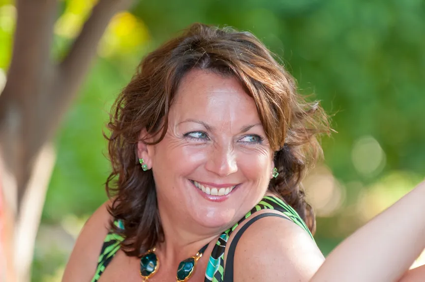 Smiling woman with brown hair wearing a green and black patterned top and green earrings, outdoors with blurred greenery background.