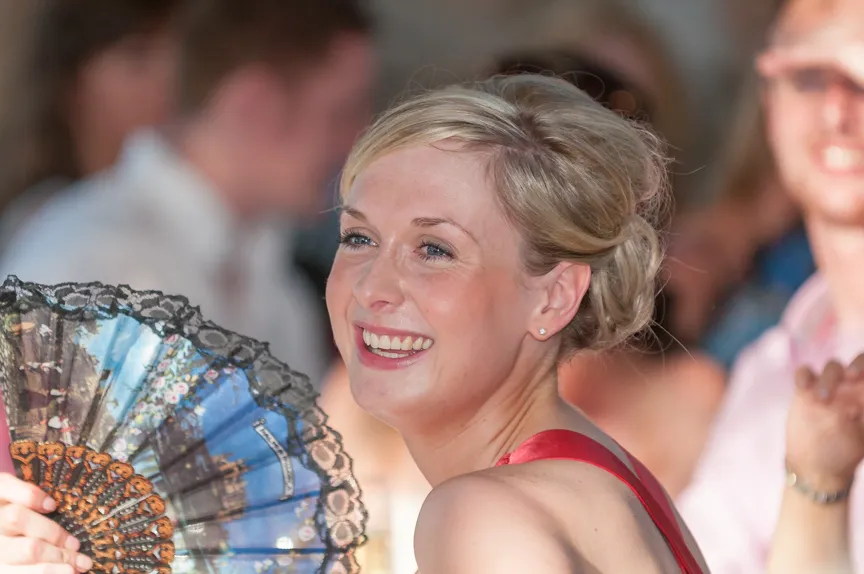 Smiling woman with blonde hair holding a decorative hand fan at an event.