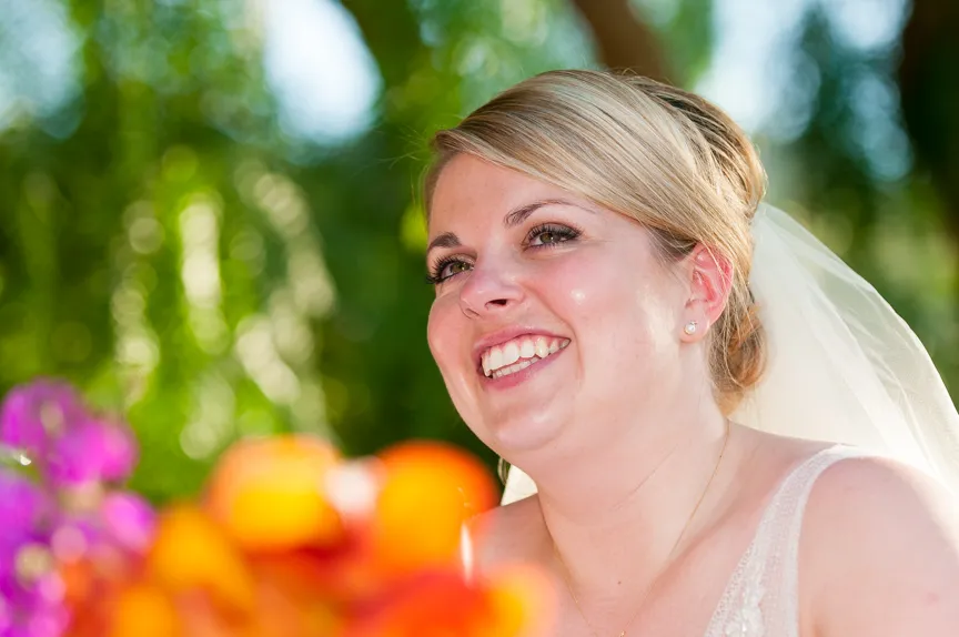 Smiling bride with blonde hair styled up and wearing a veil, with blurred colorful flowers in the foreground and green foliage background.