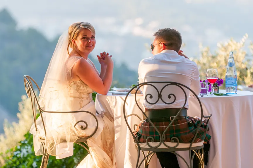 Bride in wedding dress and veil smiling at the camera sitting at an outdoor table with groom wearing a kilt and white shirt.