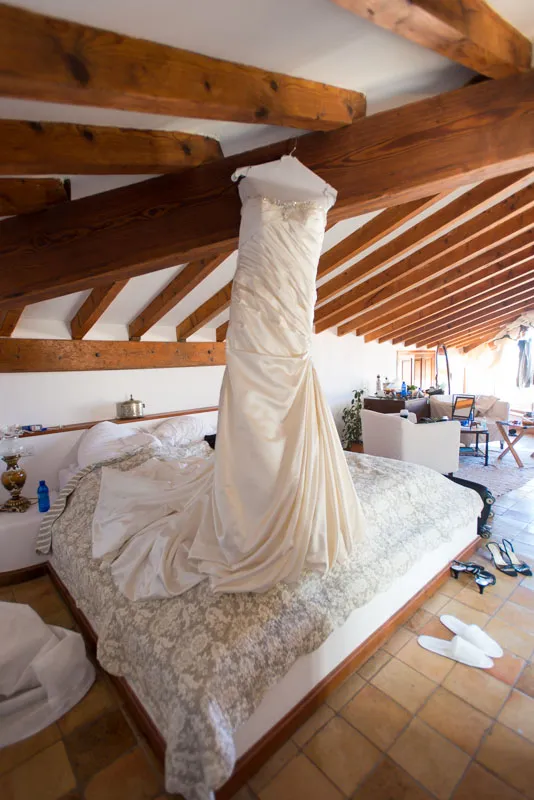 Ivory satin wedding dress hanging from wooden ceiling beams above a bed with patterned bedding in a rustic attic room.
