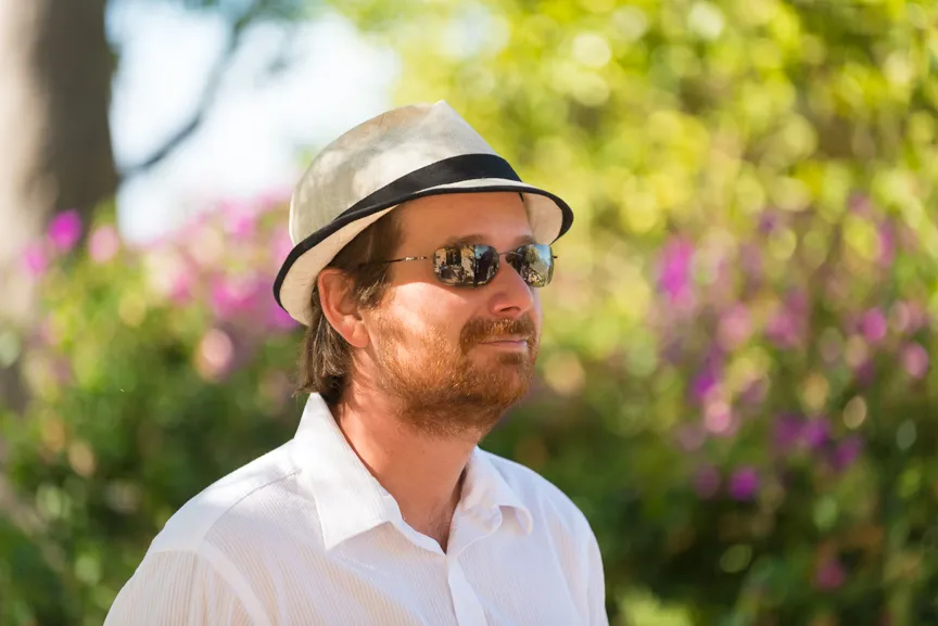 Man with a light-colored fedora hat and dark sunglasses, wearing a white shirt, standing outdoors with blurred greenery and purple flowers in the background.