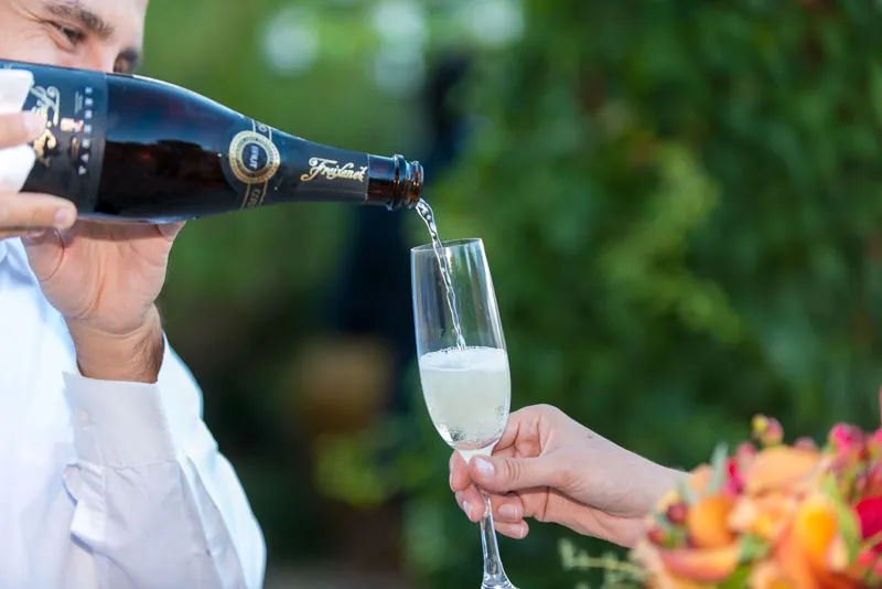 Man in white shirt pouring sparkling wine from a bottle into a champagne flute held by another person with a blurred green background.