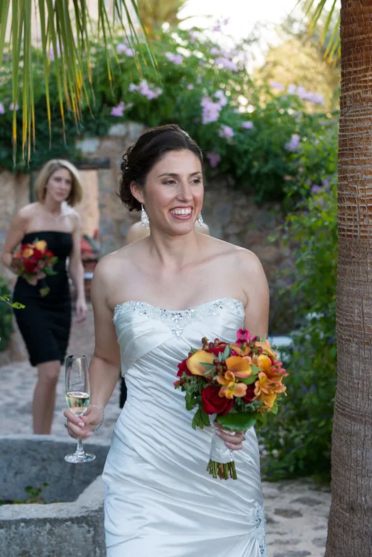 Smiling bride in white strapless wedding dress holding a bouquet of orange and red flowers and a glass of champagne outdoors.