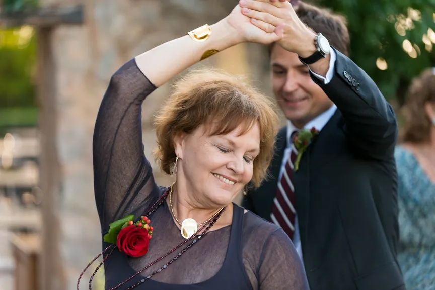 Smiling middle-aged woman with a red rose corsage dancing with a man in a suit at an outdoor event.