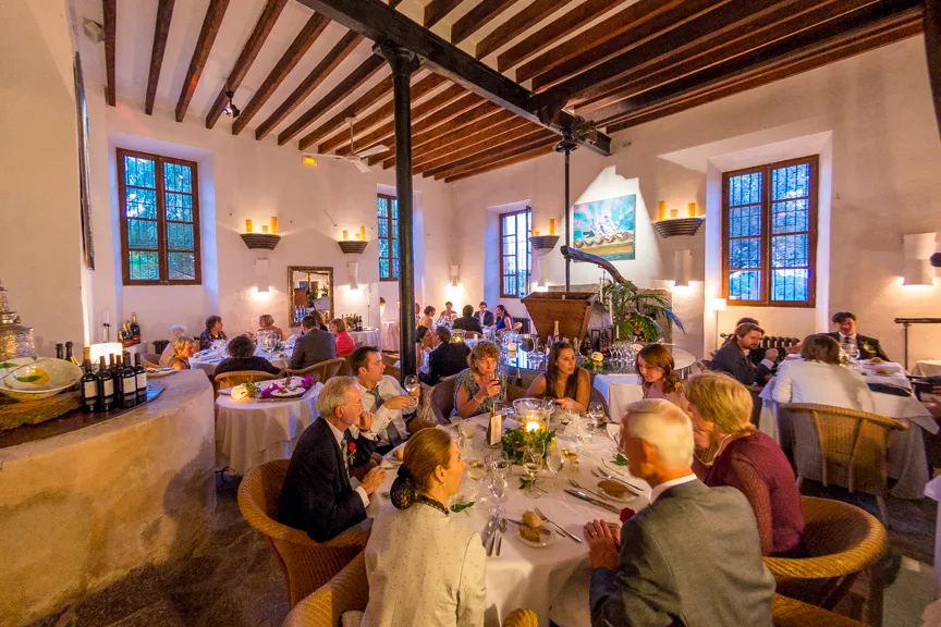 Group of people dining and socializing at round tables in a warmly lit rustic restaurant with wooden beams and large windows.