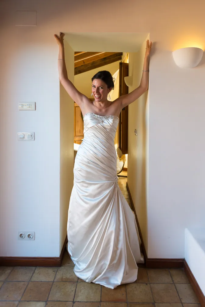 Smiling bride in a strapless white wedding gown standing in a narrow doorway with her arms raised.