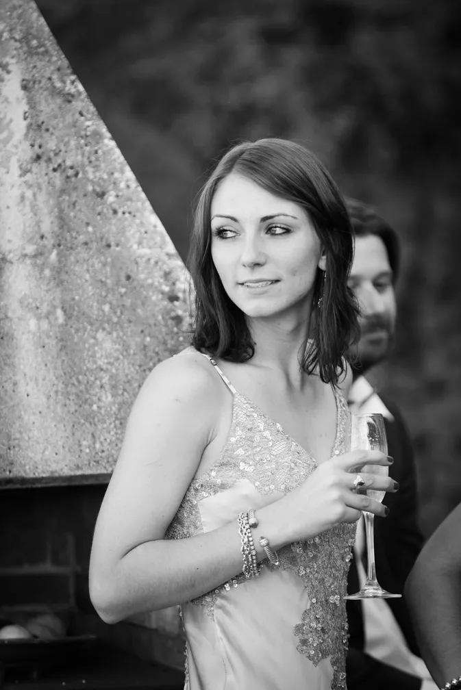 Black and white photo of a woman in a sequined dress holding a champagne glass and looking to the side, with a blurred man in the background.