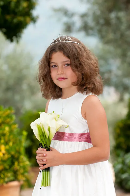 Young girl in a white dress with a pink sash wearing a tiara and holding a bouquet of white calla lilies outdoors.