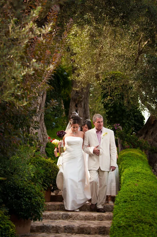 Bride in white wedding dress holding dark purple bouquet walks down stone steps arm-in-arm with an older man in a beige suit, surrounded by lush greenery.