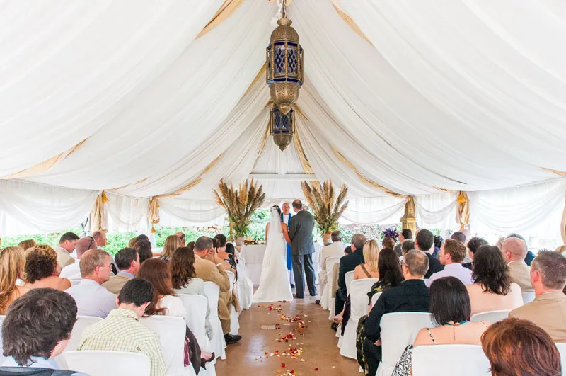 Bride and groom standing at the altar under a white draped tent with guests seated facing them during a wedding ceremony.