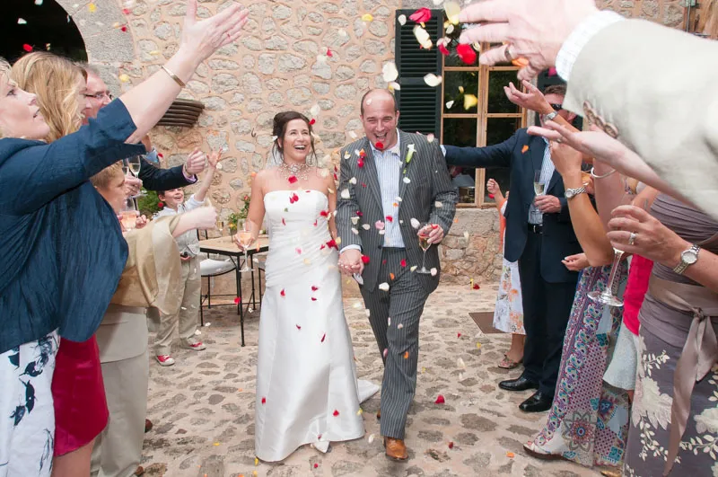 Newlywed bride and groom smiling and holding hands while guests throw rose petals around them.