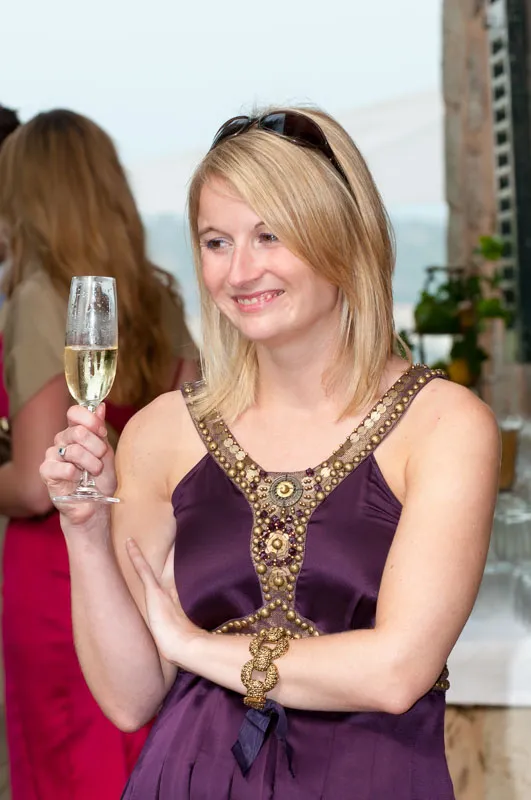 Smiling blonde woman in a purple dress holding a glass of champagne at an indoor event.