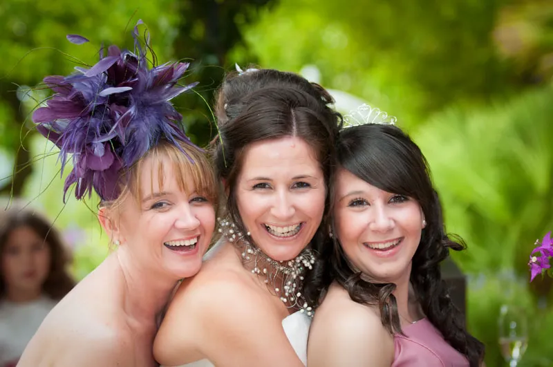 Three smiling women close together outdoors, one wearing a purple feathered headpiece and another with a tiara, likely at a wedding.