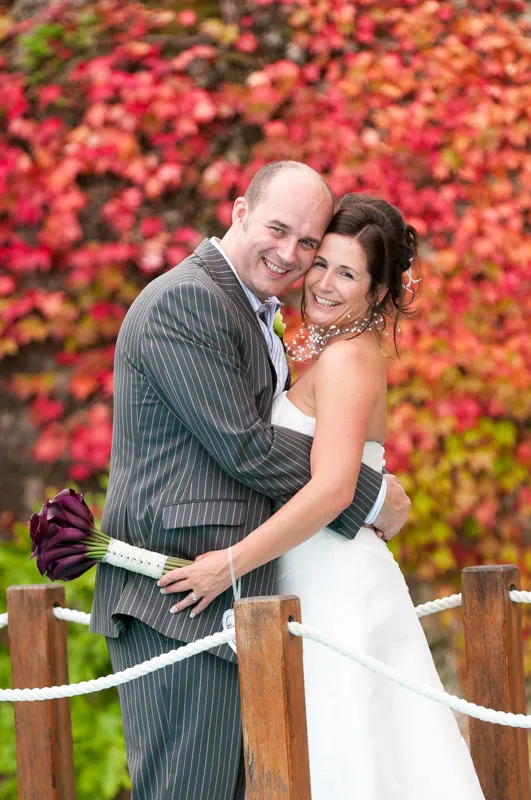 Bride and groom embracing and smiling in front of a colorful autumn foliage background.