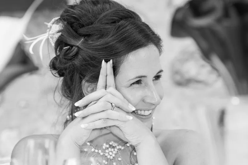 Smiling woman with styled hair resting her chin on clasped hands wearing a ring and pearl necklace.