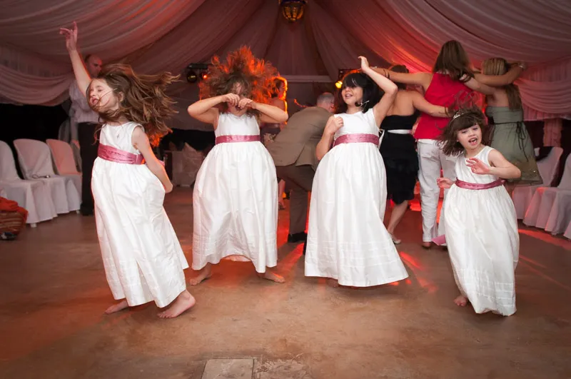 Four young girls in white dresses with pink sashes dancing energetically barefoot at an indoor event with adults in the background.