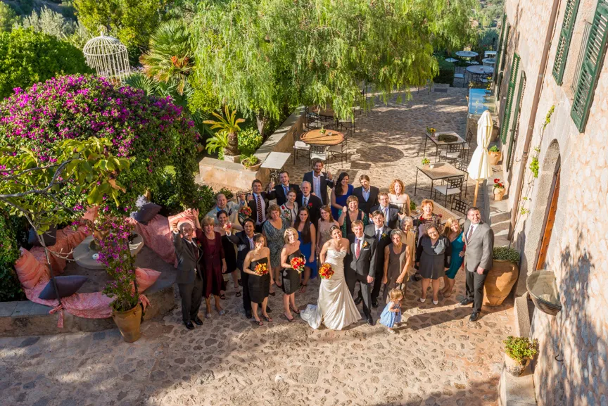 A wedding party posing outdoors on a stone patio with greenery, a pink flowering tree, and rustic building in the background.