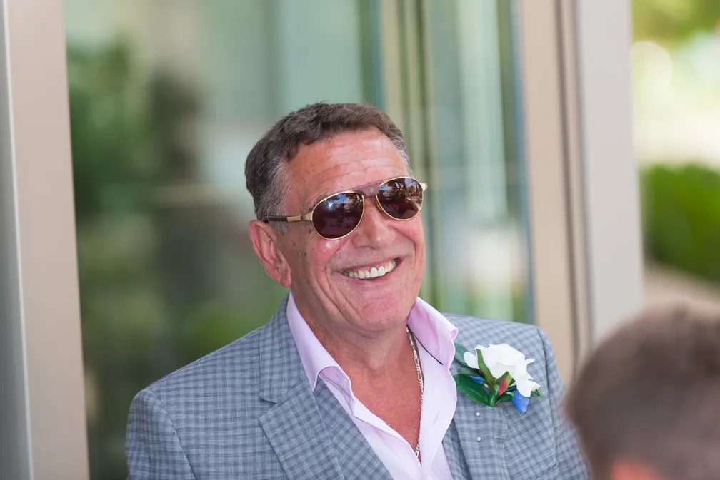 Smiling man wearing sunglasses, a pink shirt, and a gray checkered blazer with a white boutonniere.