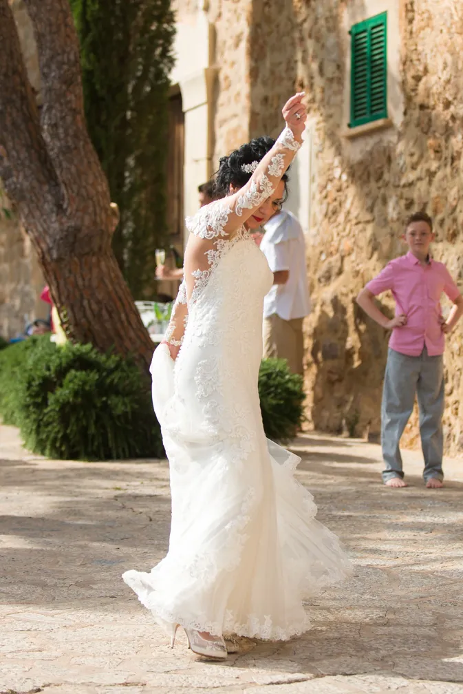 Bride in a white lace wedding dress dancing outdoors on a stone-paved area with people and a stone building in the background.