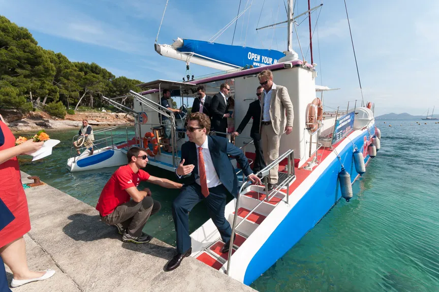 Men in suits and casual clothes boarding a blue and white sailboat at a dock near clear turquoise water and pine trees under a blue sky.