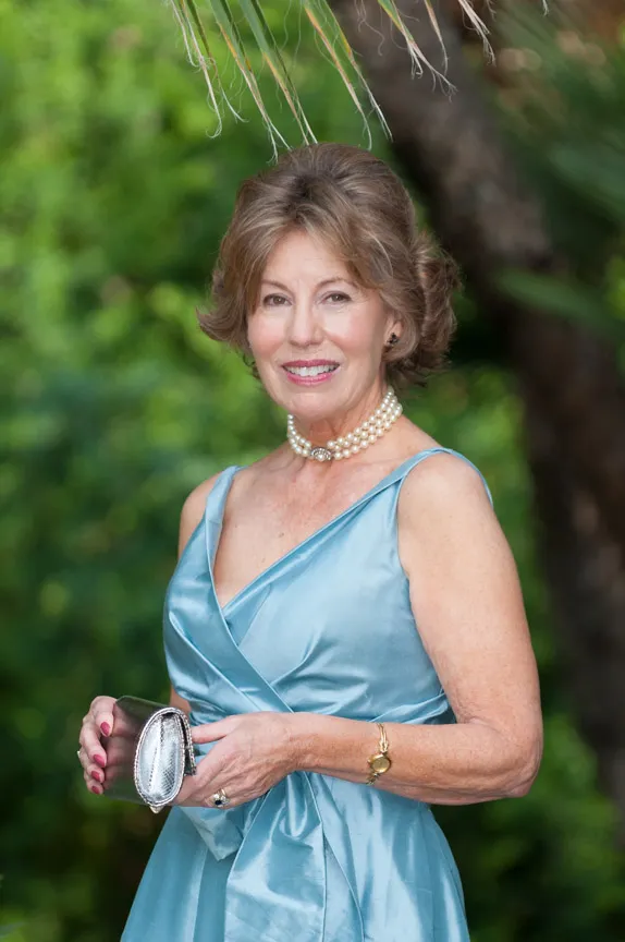Smiling older woman wearing a light blue satin dress and pearl necklace, holding a silver clutch purse outdoors.