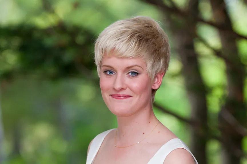 Smiling young woman with short blonde hair and blue eyes wearing a white top and a delicate necklace in an outdoor setting.