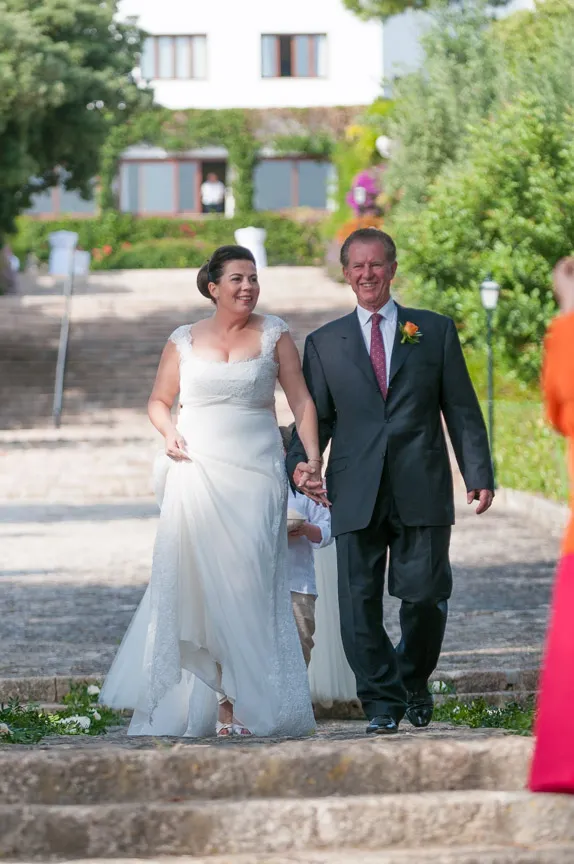 Bride in white wedding dress holding hands with a man in a black suit and red tie, walking outdoors on stone steps.