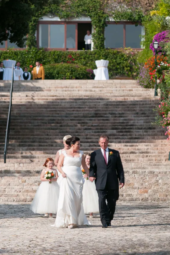 Bride in white wedding gown walking down stone steps holding hands with man in dark suit, followed by three flower girls in white dresses holding bouquets.