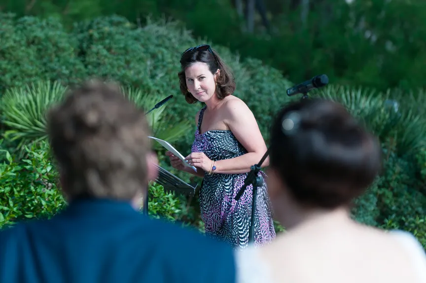 Woman in a patterned dress reading from papers at an outdoor microphone, with blurred heads of two people in the foreground.