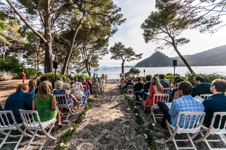 Outdoor wedding ceremony with guests seated on wooden chairs on a stone path, overlooking the sea and mountainous island in the background.