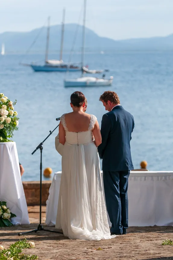 Bride in lace wedding dress and groom in blue suit standing beside a microphone near a body of water with sailboats in the background.