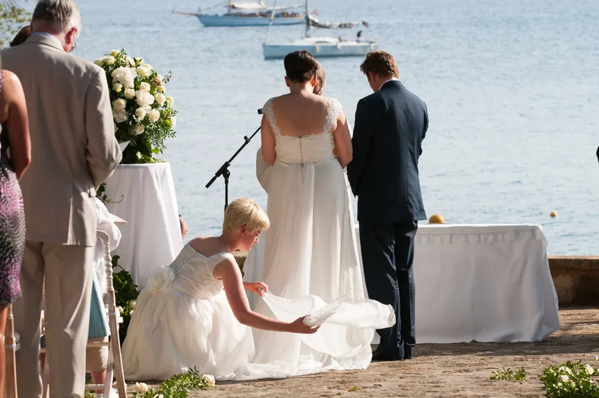Bride and groom standing by the water during an outdoor wedding ceremony, with a bridesmaid adjusting the bride’s dress.