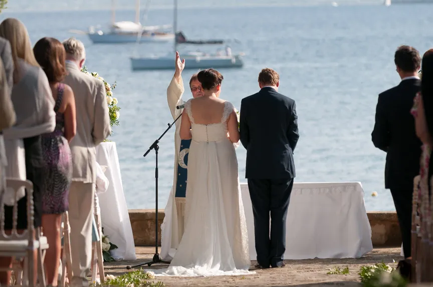 Bride and groom standing before an officiant during an outdoor waterfront wedding ceremony with guests seated on either side.