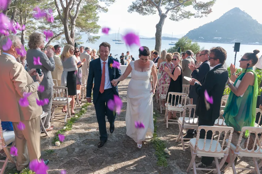 Bride and groom walking hand-in-hand down an outdoor aisle by the sea, surrounded by applauding guests and falling purple flower petals.