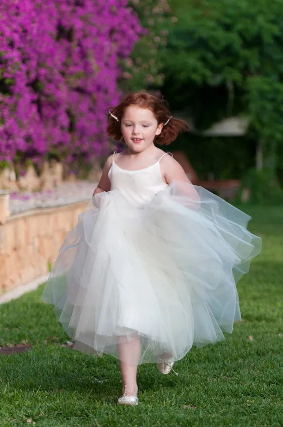 Young girl with red hair running joyfully on grass wearing a white tulle dress and ballet flats.