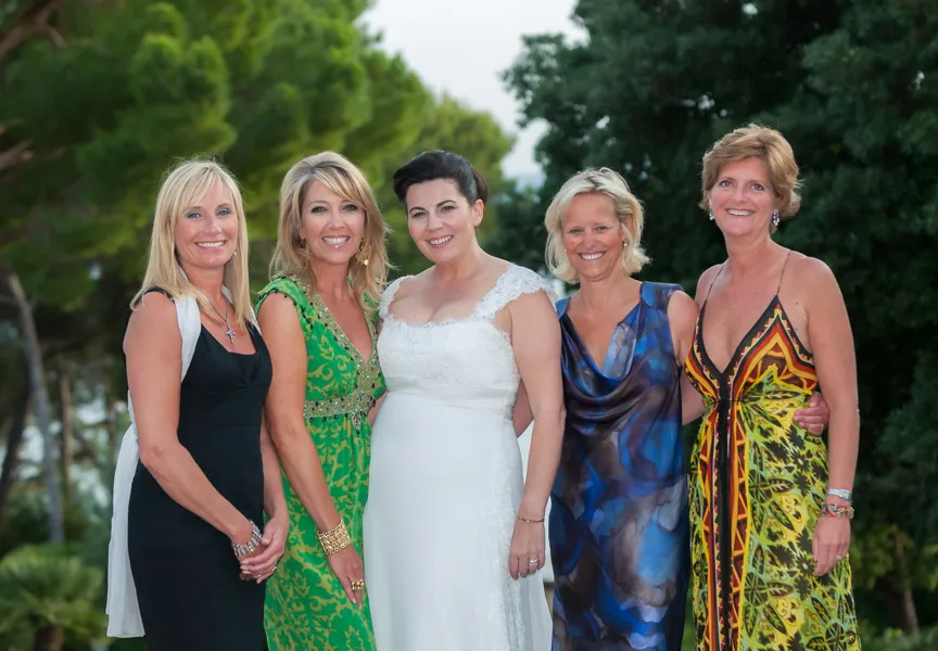 Five women standing outdoors smiling; the center woman wears a white wedding dress and the others wear colorful dresses.