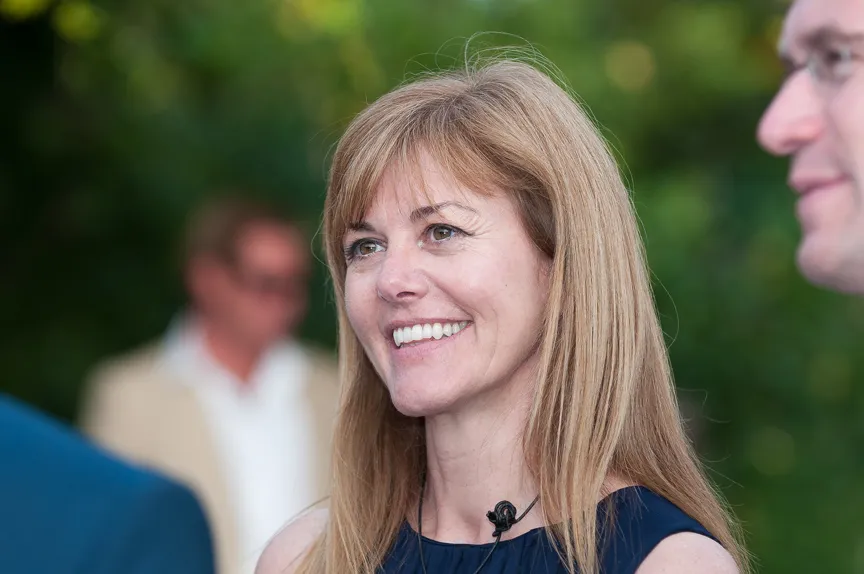 Smiling woman with long blonde hair wearing a navy blue top, talking to others at an outdoor event.