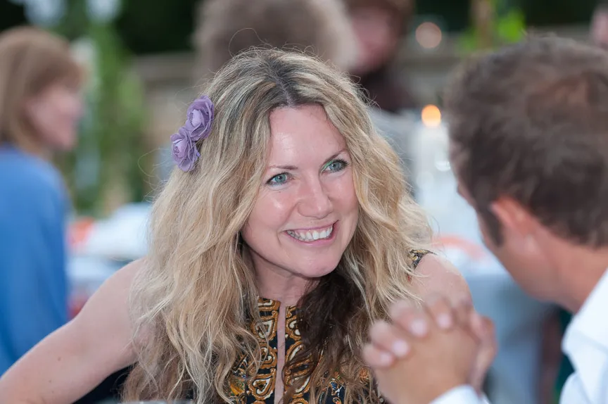 Smiling woman with long wavy blonde hair and a purple flower accessory talking to a man blurred in the foreground.