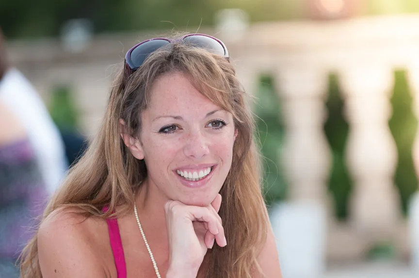 Smiling woman with long hair resting her chin on her hand outdoors, wearing sunglasses on her head and a beaded necklace.