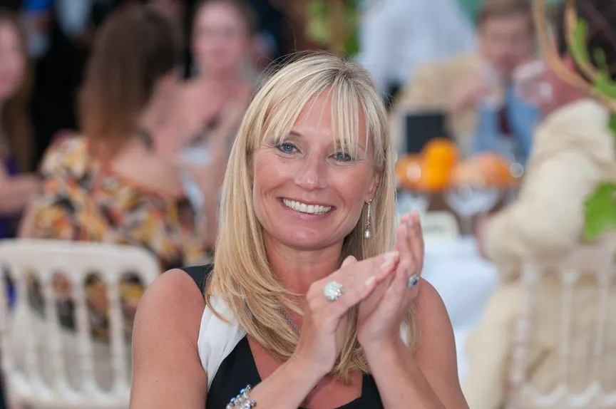Smiling blonde woman clapping hands at an indoor event with blurred people and tables in the background.