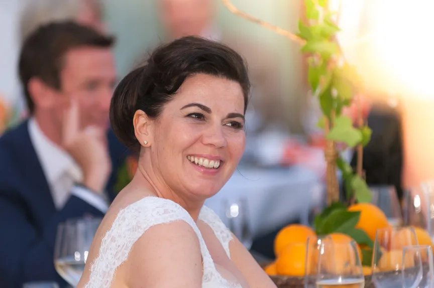 Smiling bride in a white lace dress sitting at a decorated table with oranges and wine glasses.