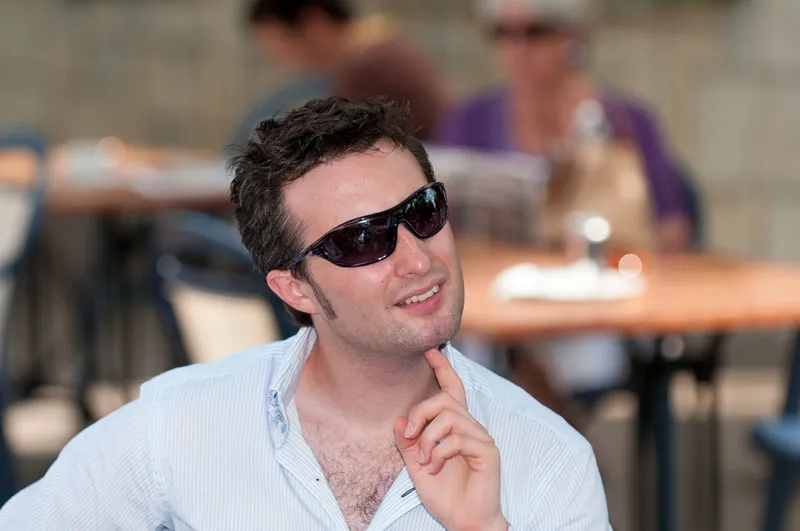 Man wearing sunglasses and a light blue striped shirt, resting his chin on his hand and looking thoughtful in an outdoor café setting.
