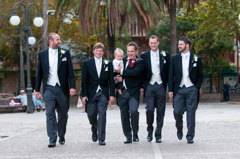 Five men in formal suits walking outdoors, one holding a baby, with trees and lampposts in the background.