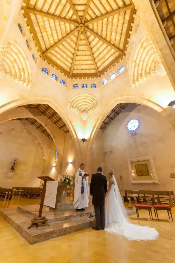 Bride and groom standing before a priest inside a church with vaulted ceiling during a wedding ceremony.