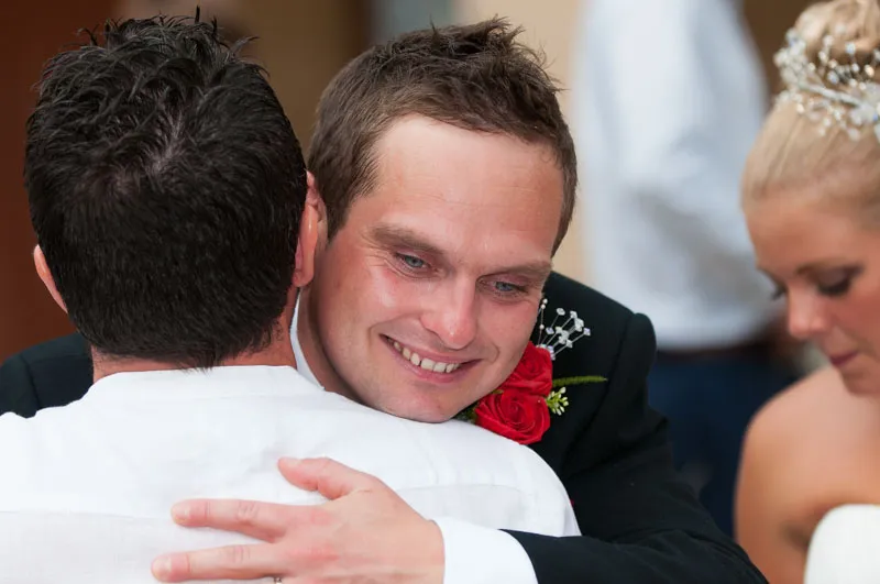 Smiling groom in a black suit with red rose boutonniere hugging a man in a white shirt, bride visible in a white dress to the right.