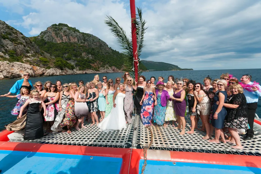 Group of women, including a bride in a white dress, posing and smiling on the netting of a colorful catamaran with rocky cliffs and ocean in the background.
